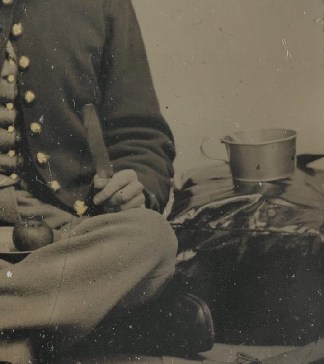 Unidentified Soldier posed in a Photographer's Tent or Studio. He has a 3-tine fork in one hand, and butter knife in the other, with an apple on his plate. But notice his shorter than "standard" tin cup, resting on top of his knapsack. 